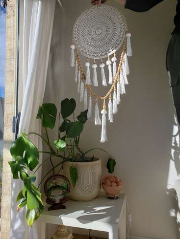 A hand holds a large, intricately woven dreamcatcher above a white table with a potted monstera plant, decorative bowl, and natural sunlight streaming in.