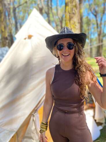 A smiling woman in sunglasses and a black hat stands outside a tent in the woods, enjoying a sunny day, dressed in a brown sleeveless outfit.
