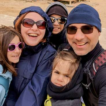 A imagem mostra uma família de cinco pessoas posando para uma selfie, vestida com roupas de inverno, com uma área de areia e um banco visíveis ao fundo.