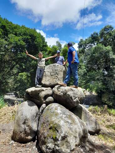 Tres personas están de pie sobre grandes rocas al aire libre. Un niño abre los brazos, sonriendo, mientras dos adultos observan. Los rodean árboles verdes, un cielo azul y la luz del sol.