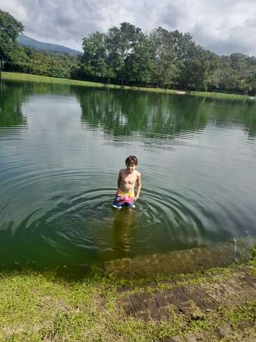 Un niño está de pie en un lago sereno, con un bañador de colores. Árboles verdes y orillas cubiertas de hierba rodean el agua, con montañas y cielos nublados al fondo.