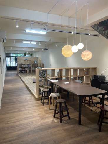 This image shows a modern, cozy library or reading area with wooden floors, tables, stools, bookshelves, hanging globe lights, and bright, inviting lighting.