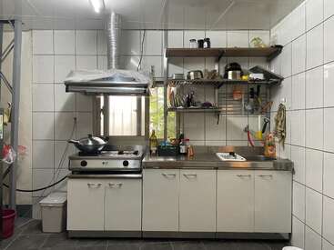 A compact kitchen with tiled walls, stainless steel countertop, gas stove, overhead exhaust, shelves with utensils, dish rack, sink, hanging towel, and cleaning supplies.