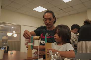 A man and a child are working together on a craft project in a classroom, focusing on building something with cardboard tubes, sticks, and string.
