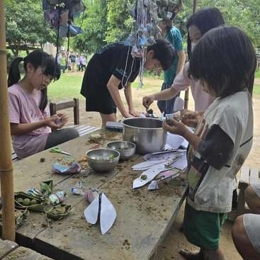 A group of children are gathered around a wooden table outdoors, crafting with leaves, papers, and bowls, deeply engaged and enjoying a creative, nature-inspired activity together.