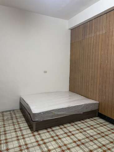 The image shows a simple, empty room with a mattress on a bed frame, plastic-wrapped, placed in the corner. The floor has patterned tiles.