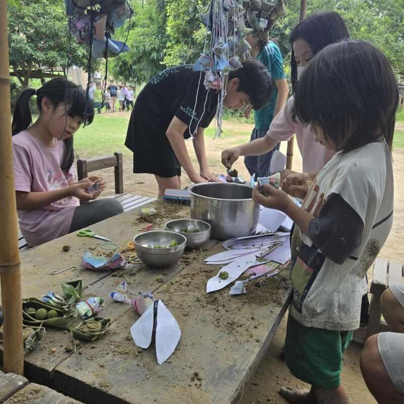A group of children are gathered around a wooden table outdoors, crafting with leaves, papers, and bowls, deeply engaged and enjoying a creative, nature-inspired activity together.