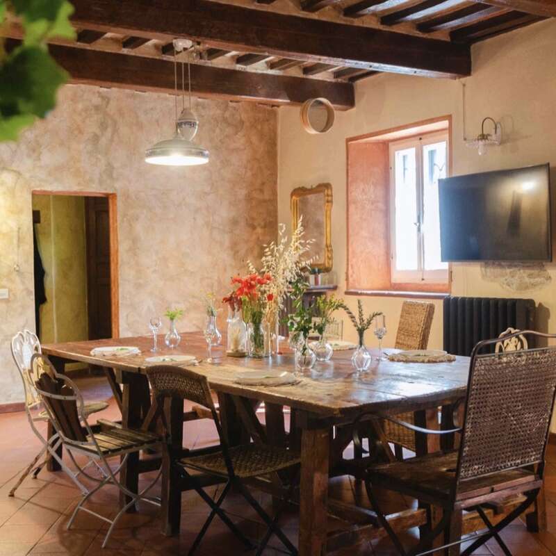 This cozy dining room features a rustic wooden table, assorted chairs, wildflower centerpieces, warm lighting, exposed beams, textured walls, a window, and mounted television.