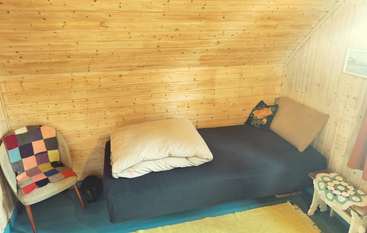 Este quarto aconchegante tem paredes com painéis de madeira, uma cama de solteiro, uma cadeira colorida, uma mesa, estantes de livros, detalhes em crochê, um tapete colorido e uma janela grande.