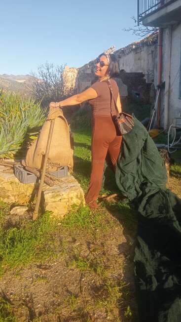 Une femme sourit en plein air, vêtue de vêtements marron et d'un bandeau, tenant un filet vert, debout près d'un sac en jute, profitant d'un environnement champêtre ensoleillé et rustique.
