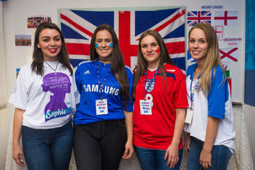 Four young women stand in front of a Union Jack flag, wearing UK-themed shirts and face paint, smiling proudly, celebrating or supporting a British event.