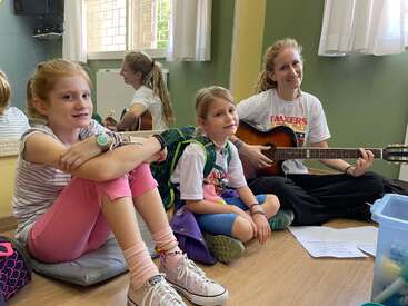 Two young girls sit on the floor, smiling, next to a woman playing guitar. They are in a music classroom with warm light and relaxed atmosphere.