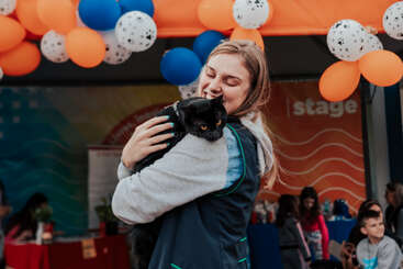 A smiling woman hugs a black cat at a festive event with colorful orange, blue, and white balloons. People and children are gathered happily in the background.