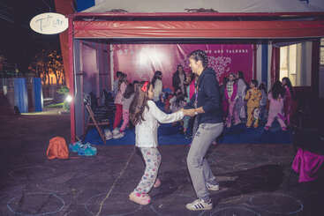 A group of children in pajamas are gathered under a tent, with two people dancing happily in the foreground. The atmosphere is joyful and playful.