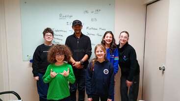 A group of five smiling kids and one adult stand together in a classroom, posing happily in front of a whiteboard with writing on it.