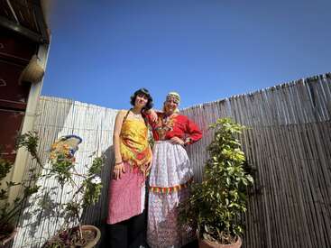 Deux femmes se tiennent côte à côte à l'extérieur, vêtues de tenues traditionnelles colorées. Elles sourient, entourées de plantes en pot devant une clôture en bambou, sous un ciel bleu clair et lumineux.