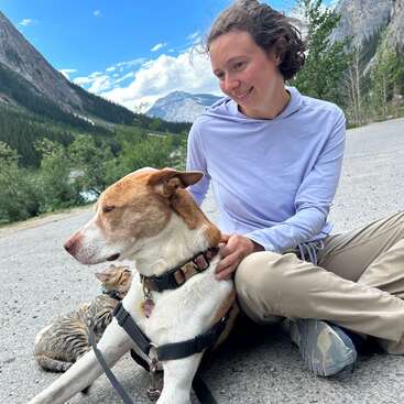 Eine Frau sitzt lächelnd mit einem Hund und einer Katze auf einer Bergstraße. Üppiges Grün und majestätische Berge rahmen die helle, friedliche Szene im Freien ein.