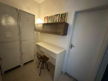Das Bild zeigt eine kleine Leseecke mit einem weißen Schreibtisch, einem Holzhocker, Metallschränken, einem Wandschrank, einer Lampe und vier bunten gerahmten Bildern darüber.