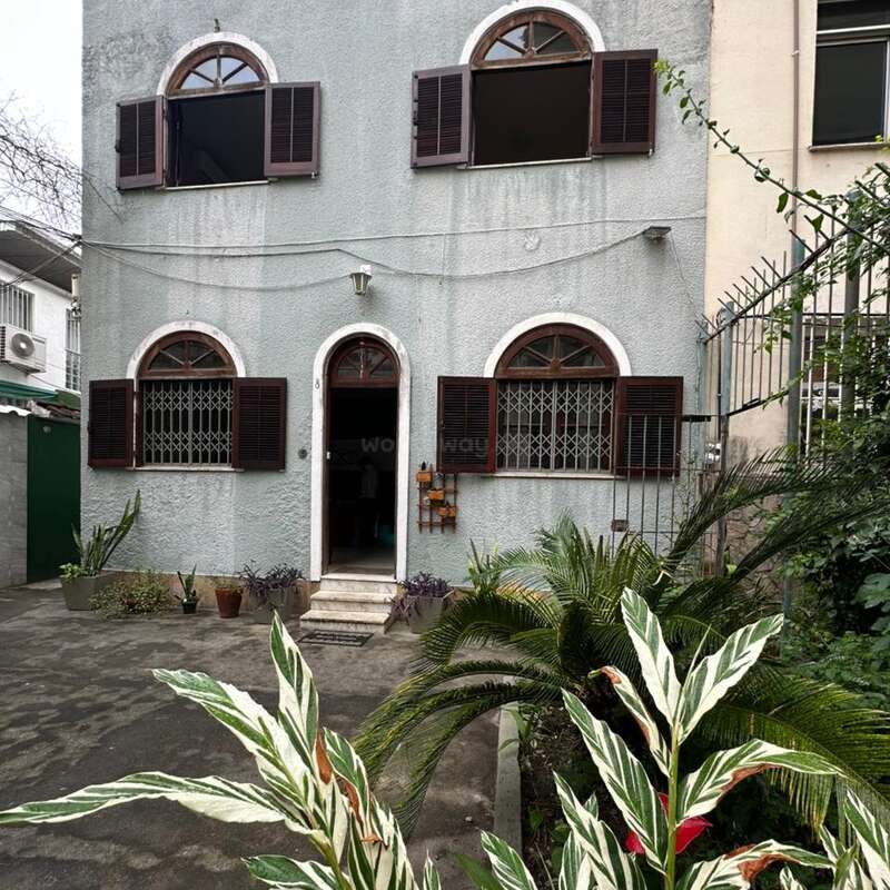 Ein zweistöckiges Haus mit Bogenfenstern, braunen Fensterläden und einer hellgrauen Fassade. Im Garten stehen Topfpflanzen, es gibt viel Grün und einen Betonweg.