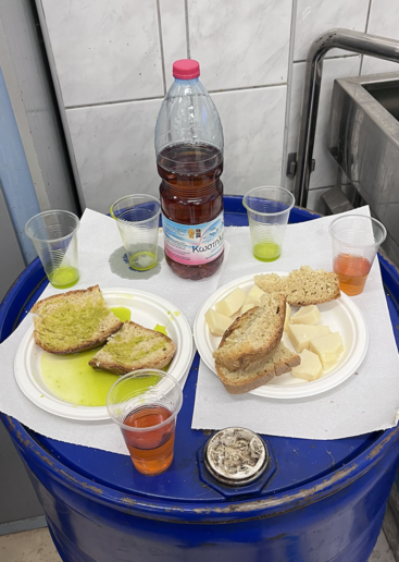 Das Bild zeigt ein blaues Fass mit einem weißen Papiertuch darauf, in dem eine Flasche Saft, zwei Teller mit Essen und vier Tassen stehen, vor dem Hintergrund einer gekachelten Wand.