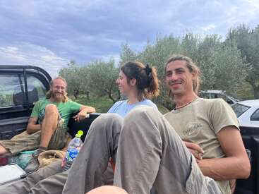 Das Bild zeigt drei Personen, die auf dem Rücksitz eines Pickups sitzen, mit Bäumen und einem bewölkten Himmel im Hintergrund, der eine entspannte Atmosphäre im Freien vermittelt.