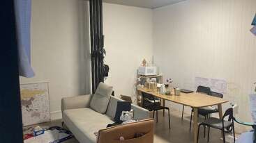 A small room has a beige sofa, a cluttered wooden table with four chairs, a microwave, maps on the wall, and some household items scattered around.