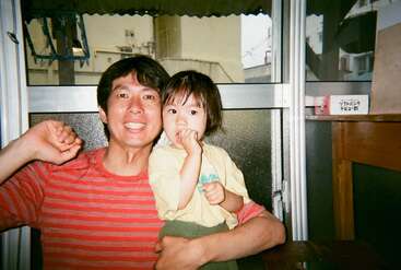 A smiling man in a red striped shirt hugs a young child in yellow indoors. They appear happy and relaxed, sitting by a large window together.