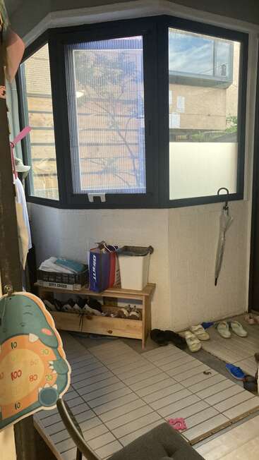 A small entryway with tiled floor, shoe rack, slippers, shopping bag, umbrella, and a dartboard. Large windows provide natural light, showing an outside building.