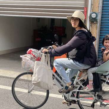 A woman wearing a sunhat rides a white bicycle with a child sitting on the back. Bags hang from the handlebars; they seem to be enjoying a sunny day.