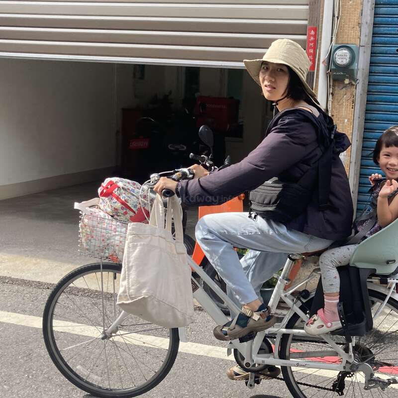 A woman wearing a sunhat rides a white bicycle with a child sitting on the back. Bags hang from the handlebars; they seem to be enjoying a sunny day.