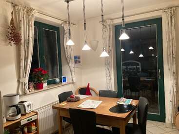 Un acogedor comedor con una mesa de madera, cuatro sillas, lámparas colgantes, cortinas con motivos florales, una ventana, una puerta, una cafetera, una planta en maceta, periódicos y un centro de mesa decorativo.