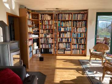 El salón, muy luminoso, cuenta con una gran estantería de madera repleta de libros, un acogedor sillón de mimbre, una estufa de leña, una puerta acristalada que da al balcón y suelo de madera.