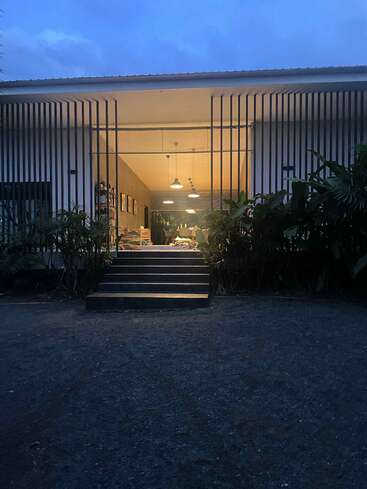 Moderno edificio iluminado al atardecer, de arquitectura abierta con lamas verticales, unos escalones de subida, rodeado de frondosas plantas y una cálida iluminación en el interior. Ambiente tranquilo y acogedor.