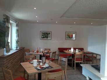 Das Bild zeigt ein Esszimmer mit weißen Wänden, Holztischen und roten Stühlen, mit Kunstwerken an den Wänden und einem Fenster mit Vorhängen.