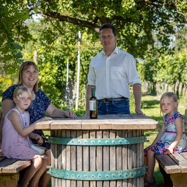 Das Bild zeigt eine vierköpfige Familie, die an einem Holztisch in einem Weinberg sitzt, mit einer Flasche Wein und Gläsern auf dem Tisch, umgeben von üppigem Grün.