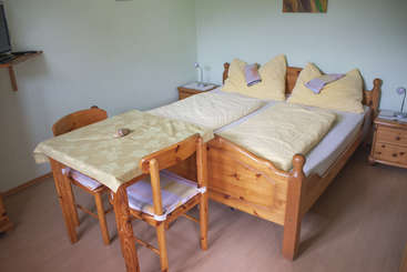 Das Bild zeigt einen Raum mit einem Bett, einem Schreibtisch und einem Stuhl, mit einem großen Fenster und einer Tür, wahrscheinlich ein Schlafzimmer oder ein Büro, mit einer neutralen Farbgebung und einem einfachen Design.