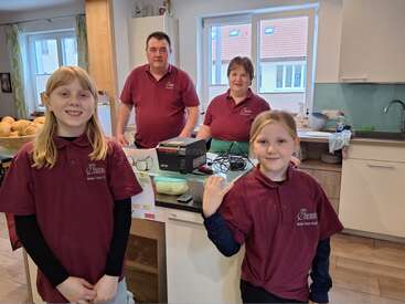 Eine vierköpfige Familie in passenden kastanienbraunen Hemden steht lächelnd in einer modernen Küche. Im Hintergrund sind Küchengeräte und Brotkörbe zu sehen.