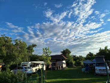 Eine friedliche Campingszene zeigt Wohnwagen, ein Auto, Zelte und einen Unterstand. Grünes Gras und Bäume stehen unter einem Himmel mit dramatischen, verstreuten Wolken.