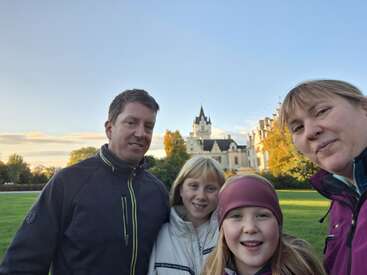 Eine vierköpfige Familie, die im Freien lächelt, steht an einem sonnigen Tag mit blauem Himmel vor einem großen Gebäude mit einem spitzen Turm.