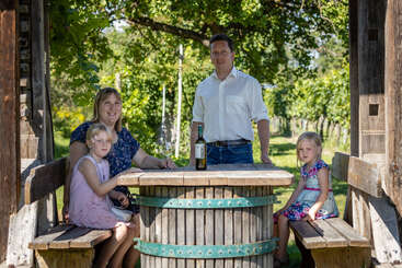 Das Bild zeigt eine vierköpfige Familie, die an einem Holztisch in einem Weinberg sitzt, mit einer Flasche Wein und Gläsern auf dem Tisch, umgeben von üppigem Grün.