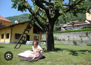 Une femme est assise sur un coussin sous un arbre dans un jardin ensoleillé. Une maison jaune, une échelle et des montagnes lointaines sont visibles à l'arrière-plan.