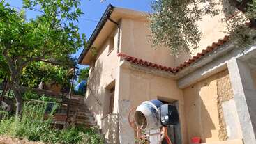 Esta imagen muestra una casa de estuco beige con tejado rojo, rodeada de árboles y vegetación. Delante hay una hormigonera, lo que sugiere que se están realizando obras.