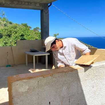 A man in a white shirt and hat works on a construction project outdoors, surrounded by lush greenery, blue sky, and ocean views in the background.