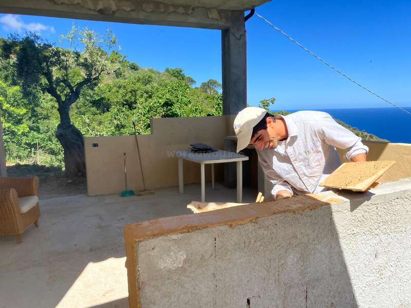 Um homem de camisa branca e chapéu trabalha em um projeto de construção ao ar livre, cercado por uma vegetação exuberante, céu azul e vista para o mar ao fundo.