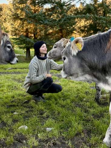 A person crouches on green grass, smiling with eyes closed, gently touching a cow’s nose, surrounded by three gray cows, with trees and sunlight in the background.