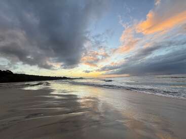 Une plage sereine au coucher du soleil, de douces vagues s'échouant sur le rivage. Des nuages spectaculaires, des lumières dorées et bleues se reflètent sur le sable mouillé, créant une scène côtière paisible, pittoresque et tranquille.