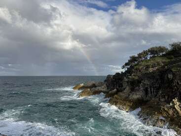 Les vagues s'écrasent contre une falaise côtière rocheuse sous un ciel nuageux dramatique. La lumière du soleil perce, mettant en évidence un léger arc-en-ciel à l'horizon, créant une atmosphère sereine.