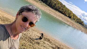 Una mujer sonriente con gafas de sol se toma un selfie a orillas de un río tranquilo, con un perro negro jugando cerca. Árboles con follaje otoñal bordean la orilla opuesta.