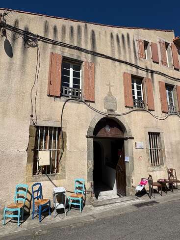 This image shows an old building with faded, rustic shutters, mismatched chairs and tables outside, wires overhead, and a partially open arched wooden door.