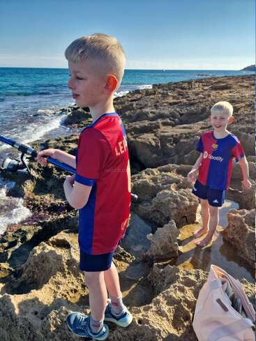 Two young boys stand on rocky shore by the sea, wearing matching Barcelona football shirts. One is fishing, the other smiling, enjoying the sunny, coastal day.